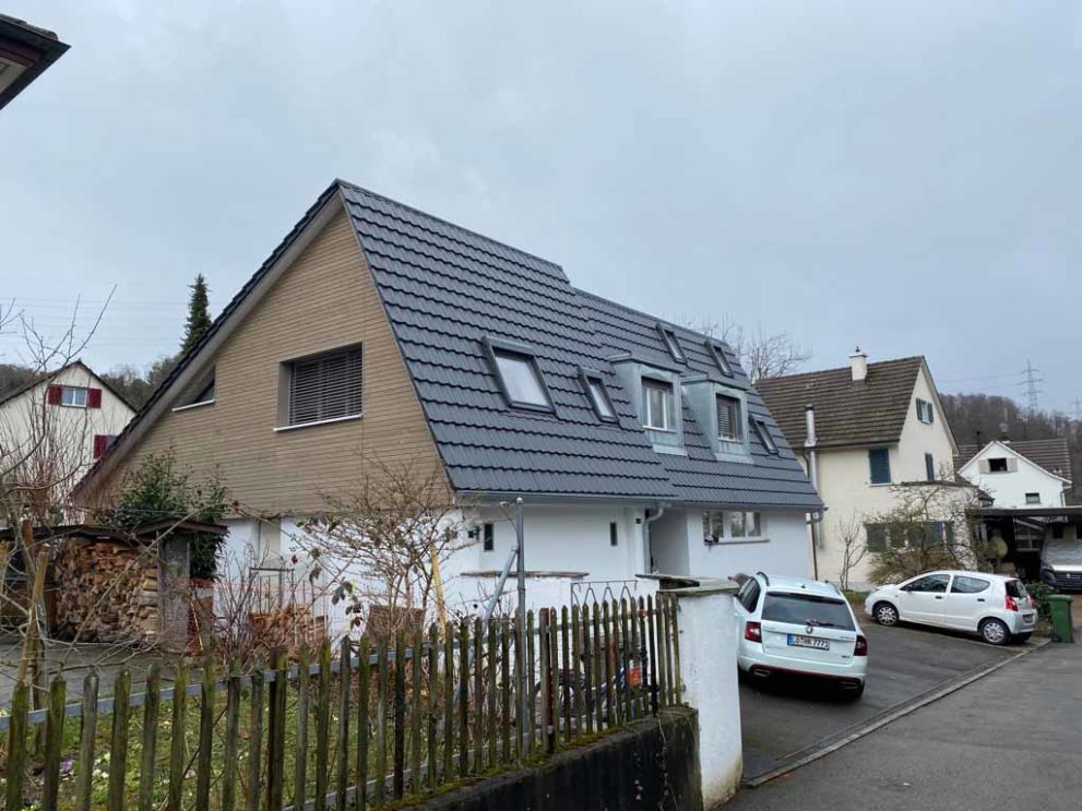 Hausansicht von der Straße, graues Steildach, weiße Fassade. Autos stehen in der Einfahrt.