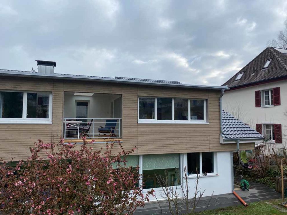 Haus mit Holzfassade und Balkon. Im Vordergrund eine Baumkrone.