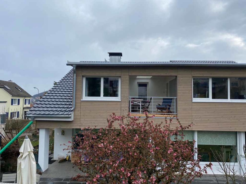 Haus mit Holzfassade und Balkon. Im Vordergrund eine Baumkrone.