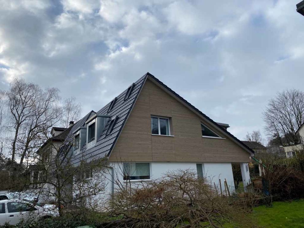 Haus mit Holzfassade ab zweitem Stock, im Vordergrund ein Garten