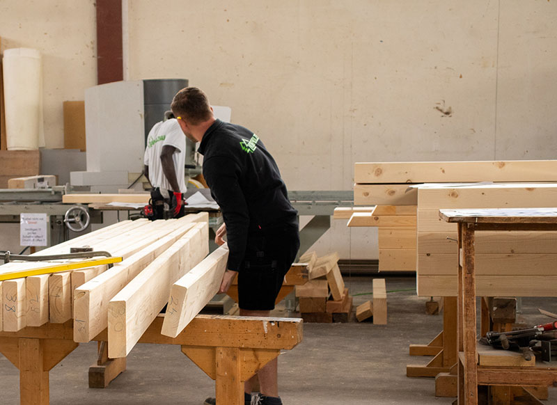 Zwei Männer in einer Arbeitshalle die lange Holzbalken bearbeiten