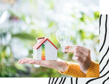Eine Hand hält ein Modellhaus mit Energieeffizienzzeichen, die andere Hand eine Glühbirne