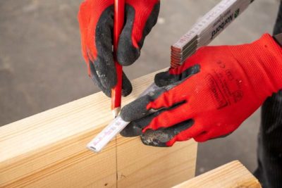 Hände in Arbeitshandschuhen, die mit Bleistift und Lineal auf einen Holzbalken zeichnen.