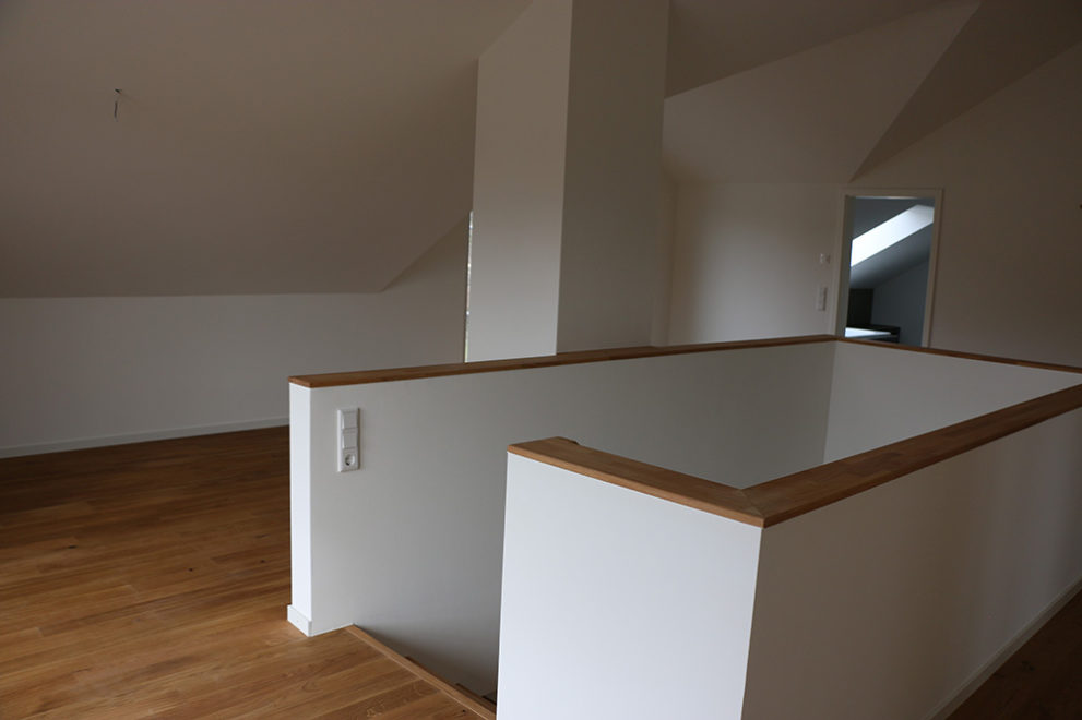 Zimmer mit Holzboden und weißen Wänden, in der Mitte des Raumes führt eine Treppe in das Erdgeschoss